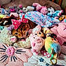 cat, orange_tabby, stuffed_animals, plush_toys, blanket, bed, bow, pink, blue, purple, cute, cozy, indoor, soft_toys, animal, sleeping, resting, childrens_room, colorful, decor