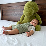 Lucas joined the competition — help win amazing prizes! baby, bed, child, comfort, cozy, cute, dinosaur, furniture, green_overalls, home, indoor, plush_toy, portrait, relaxed, resting, smiling, soft_toy, toddler, white_bed_sheet, wooden_headboard
