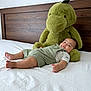 baby, bed, child, comfort, cozy, cute, dinosaur, furniture, green_overalls, home, indoor, plush_toy, portrait, relaxed, resting, smiling, soft_toy, toddler, white_bed_sheet, wooden_headboard