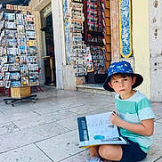 Erik is registered to the contest to win money with this photo: child, boy, hat, book, reading, street, newsstand, newspapers, postcards, tiles, building, outdoor, casual_clothing, sitting, summer, daylight, curious, young, urban, sidewalk