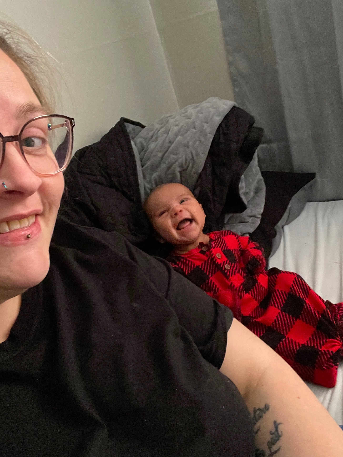 Zayden is registered to the contest to win money with this photo: adult, baby, bed, blanket, black_clothing, checkered_pattern, curtain, eyewear, facial_piercing, glasses, happy, indoor, laughing, person, plaid, portrait, smile, tattoo, toddler, wearing