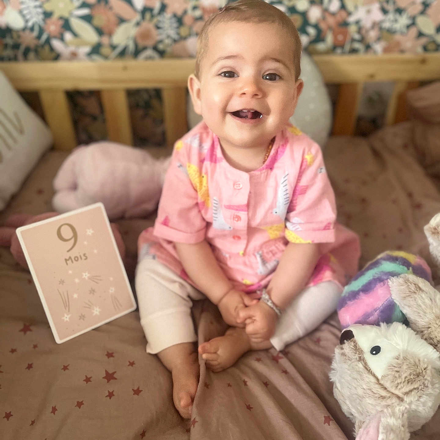 Edène a rejoint le concours — aidez-le/la à gagner de superbes lots ! baby, bed, blanket, child, cozy, cute, decor, face, happy, indoor, infant, milestone_card, pink_clothing, plush_toy, portrait, sitting, smiling, star_pattern, toy, wooden_bedframe