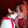 cat, woman, person, pet, portrait, selfie, kiss, affection, ribbon, pink_ribbon, collar, bell, whiskers, white_fur, tabby, red_clothing, tattoo, indoor, closeup, humor