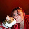 cat, woman, person, pet, kissing, affection, portrait, selfie, indoor, red_lighting, red_clothing, whiskers, collar, bell, feline, face, close_up, companion_animal, cuddle, photography
