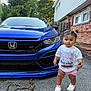 toddler, child, car, blue_car, honda, si_badge, sidewalk, house, brick_wall, trees, outdoor, daylight, concrete, stick, white_shoes, pink_shorts, white_tshirt, pigtails, curly_hair, portrait