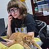 person, glasses, cat, orange_cat, sleeping_cat, indoor, cushion, yellow, black_clothing, phone, hair, furniture, cabinet, container, relaxing, hand, face, room, casual, comfort