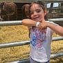 Ohana participe au concours pour gagner de l'argent avec cette photo : child, girl, smiling, outdoor, fence, cows, animal, hay, wristband, cartoon_clothing, shorts, tshirt, sunlight, tree, nature, playful, standing, person, daytime, farm
