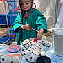 child, toy, stethoscope, stuffed_animal, play, doctor, green_gown, medical_table, syringe, tent, outdoor, smiling, pretend_play, young_girl, blue_mesh_hanging, plastic_container, white_container, play_kitchen, spot_pattern, happy