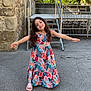 child, girl, dress, colorful, long_hair, smiling, outdoor, stone_wall, metal_stairs, pavement, sandals, sunny, greenery, tree, fence, building, happy, arms_outstretched, daylight, portrait