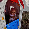 toddler, child, playhouse, outdoor, smiling, happy, plastic, toy, door, red, blue, grass, leaves, fence, daylight, reaching, young_child, person, play, fun