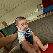 Aiden Thon joined the competition — help win amazing prizes! baby, child, indoor, carpet, person, clothing, vest, shorts, sitting, floor, bench, waiting_area, curious_expression, hair, skin, hand, foot, modern, room, lighting