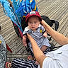 baby, child, person, cap, red_cap, adult, hands, stroller, bag, blue_bag, boardwalk, wooden_floor, outdoor, casual_clothing, striped_pants, flip_flops, seated, summer, daylight, expression