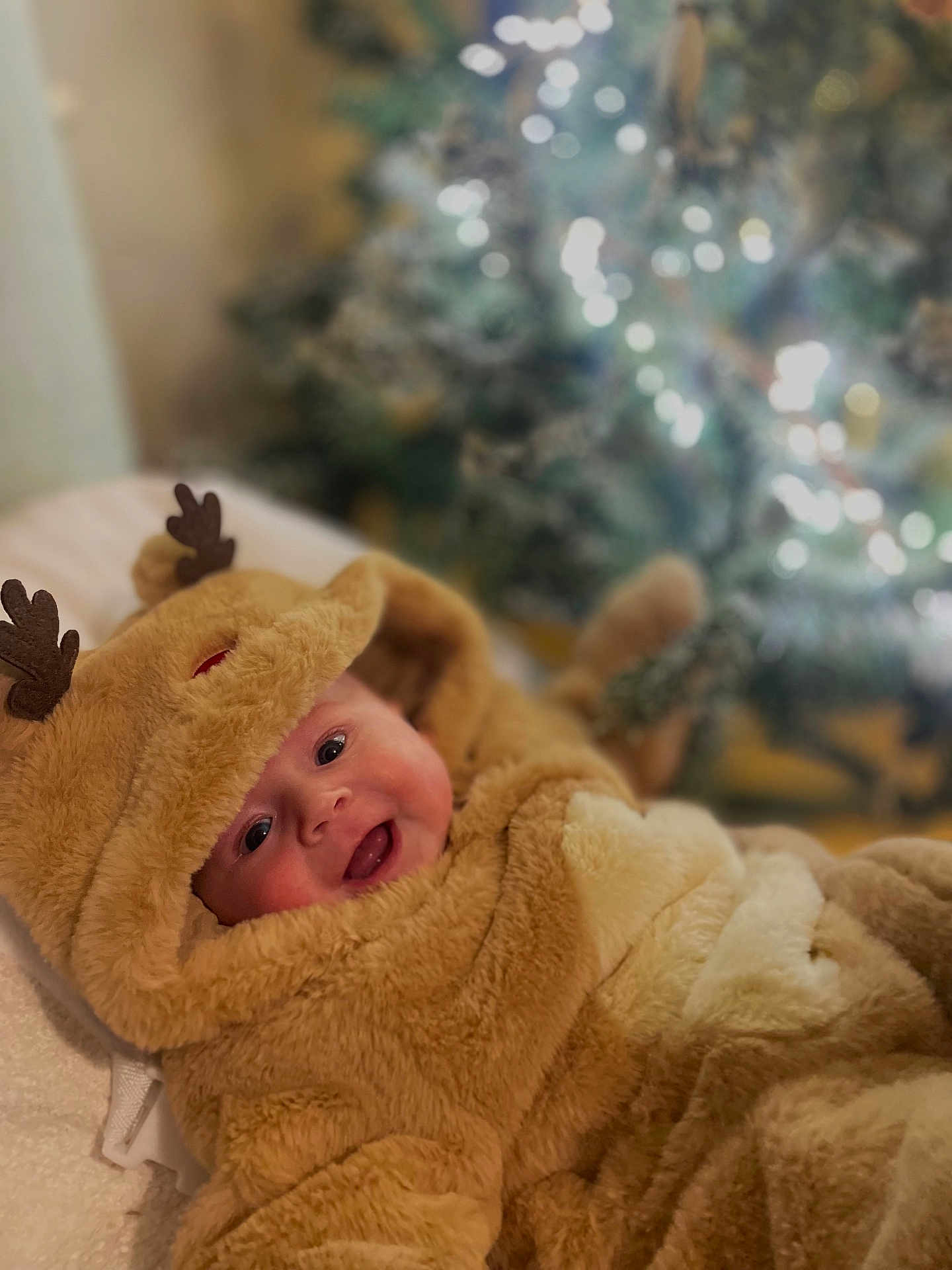 Rosie is registered to the contest to win money with this photo: baby, child, costume, reindeer, smile, face, cute, holiday, christmas_tree, blurred_background, soft_texture, warm_lighting, indoors, festive, hood, fur, lying_down, happy, infant, seasonal