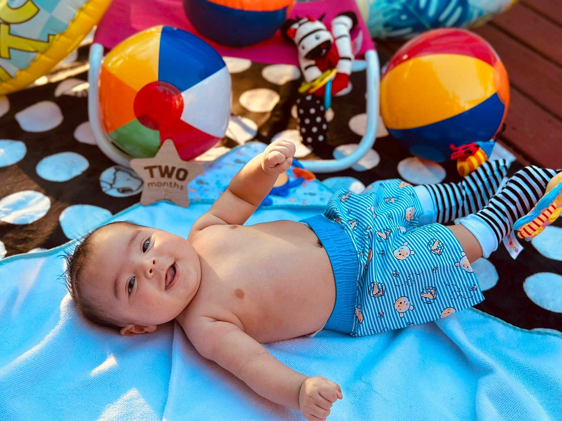 Anthony is registered to the contest to win money with this photo: baby, infant, two_months, blanket, beach_ball, toy, play_mat, smiling, shirtless, shorts, striped_socks, outdoor, wooden_deck, colorful, child, happy, cute, lying_down, playtime, sunlight