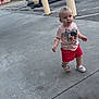 toddler, child, sidewalk, concrete, car, bollards, trees, outdoor, casual_clothing, shorts, tshirt, footwear, parking_lot, daylight, person, walking, urban, curious, young_child, background_building