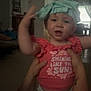 baby, child, person, indoor, clothing, shirt, hand, arm, face, expression, smile, curtain, window, furniture, living_room, adult_hand, headwear, text_on_clothing, playful, home