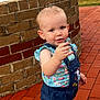 toddler, child, baby, brick_wall, brick_path, outdoor, grass, people, holding_object, blue_clothing, patterned_shirt, shorts, earring, curly_hair, daylight, park, casual, walking, smiling, portrait
