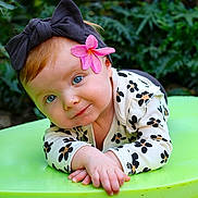 Evelynn joined the competition — help win amazing prizes! baby, child, headband, flower, blue_eyes, green_background, leafy_plants, long_sleeve, patterned_clothing, cute, portrait, infant, outdoor, nature, smiling, face, hands, lying_down, curious, closeup