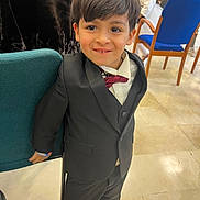 Chiheb a rejoint le concours — aidez-le/la à gagner de superbes lots ! child, boy, smile, suit, bow_tie, indoors, chair, people, floor, tile_floor, formal_clothing, hair, face, person, event, background, jacket, pants, shoes, happy