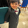 child, boy, smile, suit, bow_tie, indoors, chair, people, floor, tile_floor, formal_clothing, hair, face, person, event, background, jacket, pants, shoes, happy