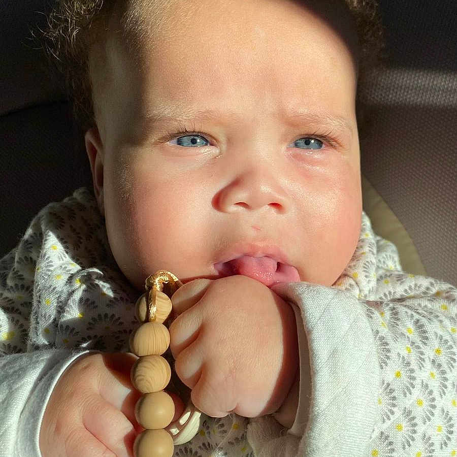 Athena a rejoint le concours — aidez-le/la à gagner de superbes lots ! adorable, baby, blue_eyes, child, close_up, cozy_clothing, curly_hair, cute, expression, face, hand, indoors, infant, pacifier_clip, patterned_outfit, person, skin, sunlight, wooden_beads, young