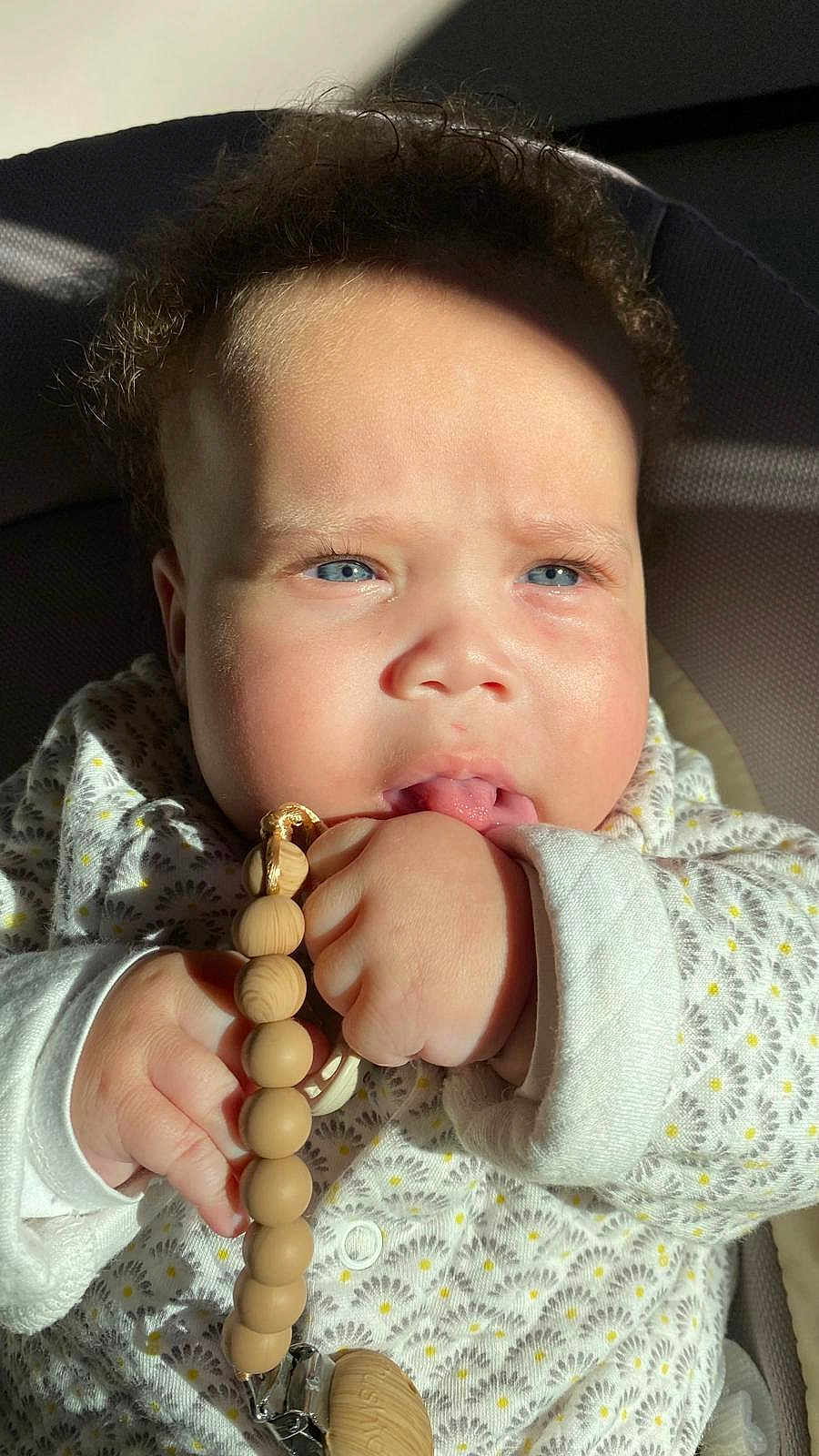 Athena a rejoint le concours — aidez-le/la à gagner de superbes lots ! adorable, baby, blue_eyes, child, close_up, cozy_clothing, curly_hair, cute, expression, face, hand, indoors, infant, pacifier_clip, patterned_outfit, person, skin, sunlight, wooden_beads, young