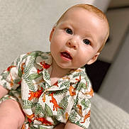 Elijah is registered to the contest to win money with this photo: baby, child, infant, dinosaur_shirt, patterned_clothing, curious_expression, blue_eyes, red_hair, sitting, soft_surface, indoor, portrait, closeup, short_sleeves, young_child, cute, face, skin, person, home