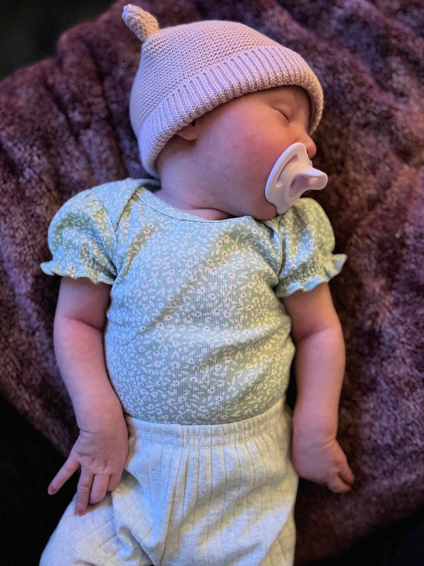 Maive is registered to the contest to win money with this photo: baby, sleeping, knitted_hat, pacifier, green_shirt, white_pants, blanket, plush, cozy, infant, child, resting, soft_lighting, portrait, cute, clothing, hand, face, headwear, relaxation