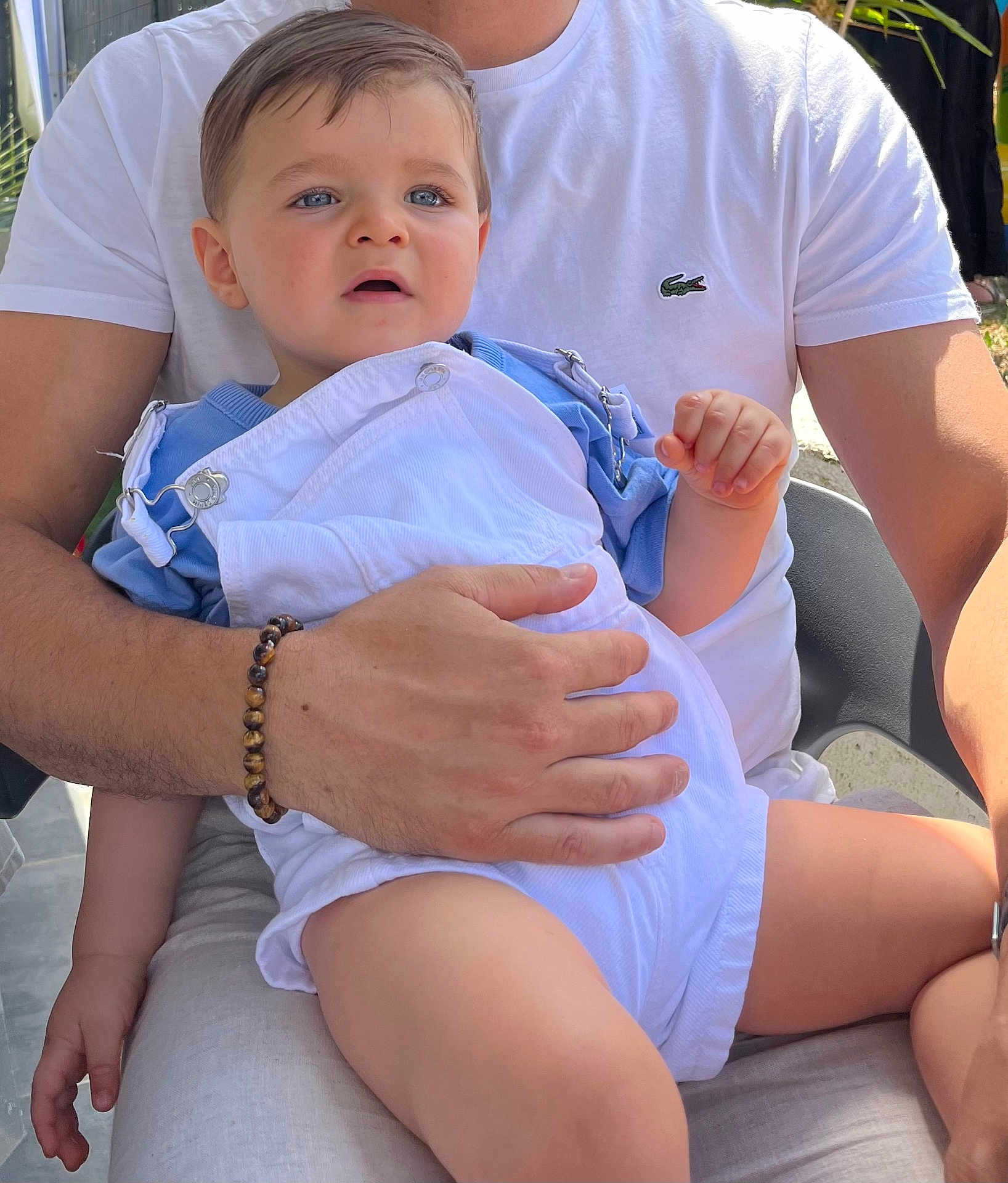 Adélio participe au concours pour gagner de l'argent avec cette photo : toddler, child, adult, blue_eyes, white_overalls, blue_shirt, bracelet, hand, lap, outdoor, sunlight, person, casual_clothing, sitting, face, portrait, expression, skin, short_hair, comfort