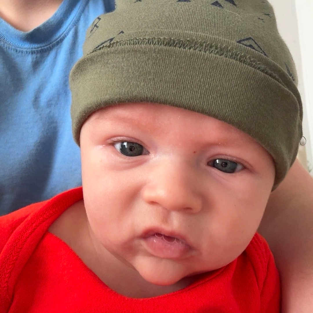 Grayson joined the competition — help win amazing prizes! baby, beanie, blue_shirt, casual_clothing, child, closeup, curious, expression, face, hat, headwear, holding, human, indoor, infant, person, portrait, red_shirt, skin, young_child