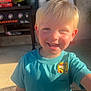 child, smiling, blonde_hair, teal_shirt, sticker, outdoor, porch, grill, sunlight, happy, face, person, casual_clothing, daylight, young_child, cute, portrait, boy, short_sleeves, background