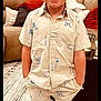 child, boy, blonde_hair, smiling, standing, shorts, shirt, blue_shoes, living_room, couch, pillow, carpet, decor, window_blinds, hands_in_pockets, patterned_clothing, indoor, happy, person, casual