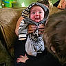 baby, child, adult, hoodie, striped_clothing, couch, plush_toy, green_toy, hand, living_room, portrait, smile, person, family, photo_frame, curly_hair, furniture, casual_clothing, indoor, holding