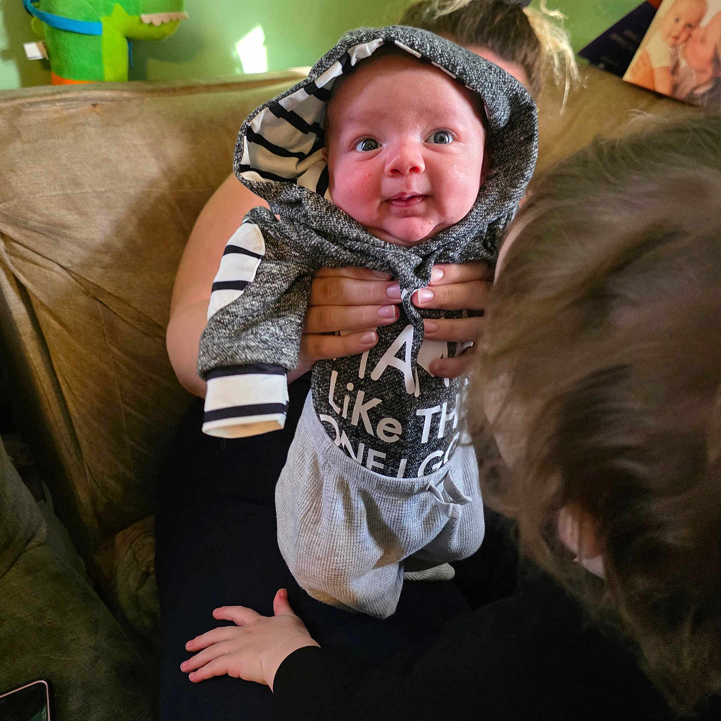 Samuel joined the competition — help win amazing prizes! baby, child, adult, hoodie, striped_clothing, couch, plush_toy, green_toy, hand, living_room, portrait, smile, person, family, photo_frame, curly_hair, furniture, casual_clothing, indoor, holding