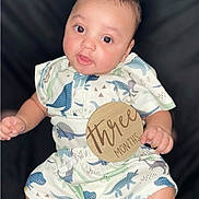 Kyrie joined the competition — help win amazing prizes! baby, infant, child, three_months, dinosaur_outfit, cute, portrait, holding_sign, wooden_sign, clothing, sitting, smiling, face, skin, hands, eyes, dark_background, young_child, adorable, indoor