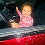 child, toddler, car, steering_wheel, interior, seat, clothing, plaid_shirt, curly_hair, smiling, hand, dashboard, window, red_car, seat_belt, person, automobile, happy, indoors, portrait