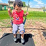 child, toddler, swing, playground, outdoor, grass, blue_sky, clouds, curly_hair, red_shirt, shorts, black_shoes, chains, sunny_day, smiling, holding, park, recreation, casual_clothing, happy