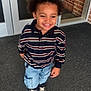 child, smiling, striped_shirt, denim_shorts, sneakers, curly_hair, happy, standing, door, glass, carpet, brick_wall, casual_clothing, young, portrait, outside, playful, cute, person, daylight