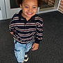 child, smiling, boy, striped_shirt, cargo_pants, sneakers, indoor, carpet, door, glass, brick_wall, happy, walking, casual_clothing, portrait, face, hair, young, person, daylight