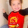 child, smiling, red_shirt, lightning_mcqueen, cartoon_car, curly_hair, happy, portrait, indoor, young_child, casual_clothing, cute, person, face, expression, joy, toddler, bright, colorful, closeup