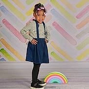 Mileena is registered to the contest to win money with this photo: child, girl, smiling, standing, striped_shirt, blue_skirt, black_tights, black_shoes, hair_beads, hair_clips, colorful_background, pastel_colors, rainbow_prop, wooden_floor, studio_photo, pose, hands_on_hips, indoor, happy, portrait