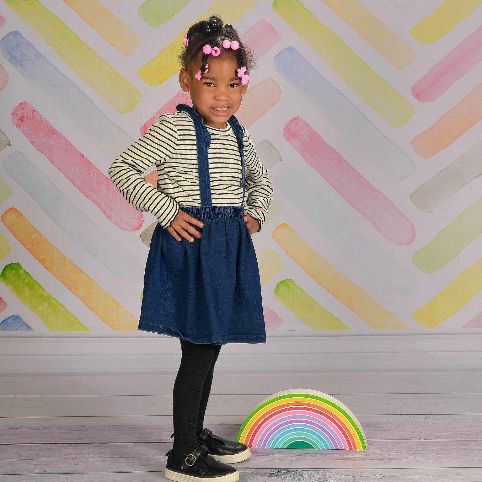 Mileena is registered to the contest to win money with this photo: black_shoes, black_tights, blue_skirt, child, colorful_background, girl, hair_beads, hair_clips, hands_on_hips, happy, indoor, pastel_colors, portrait, pose, rainbow_prop, smiling, standing, striped_shirt, studio_photo, wooden_floor