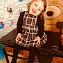 Lyna participe au concours pour gagner de l'argent avec cette photo : toddler, child, smiling, plaid_dress, black_tights, curly_hair, table, indoor, toy_bear, wallpaper, animated_characters, happy, cute, portrait, seated, playful, cozy, childhood, room, furniture