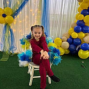 Lou a rejoint le concours — aidez-le/la à gagner de superbes lots ! child, girl, pigtails, chair, balloons, party_decor, tulle, fairy_lights, smile, crossed_legs, indoor, celebration, red_outfit, black_socks, green_floor, white_chair, blue_tulle, yellow_balloons, happy, portrait