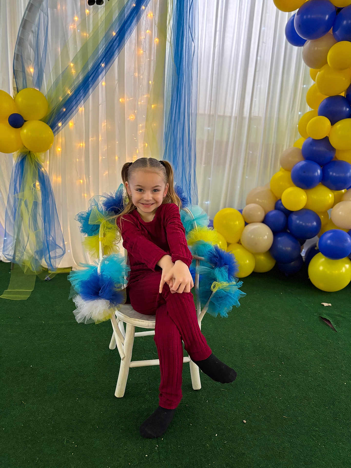Lou a rejoint le concours — aidez-le/la à gagner de superbes lots ! child, girl, pigtails, chair, balloons, party_decor, tulle, fairy_lights, smile, crossed_legs, indoor, celebration, red_outfit, black_socks, green_floor, white_chair, blue_tulle, yellow_balloons, happy, portrait