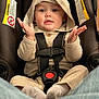 baby, child, car_seat, hooded_sweater, beige_clothing, safety_harness, infant, cute, hands_up, blue_eyes, warning_label, indoor, seatbelt, curious_expression, soft_fabric, casual, person, portrait, young, sitting