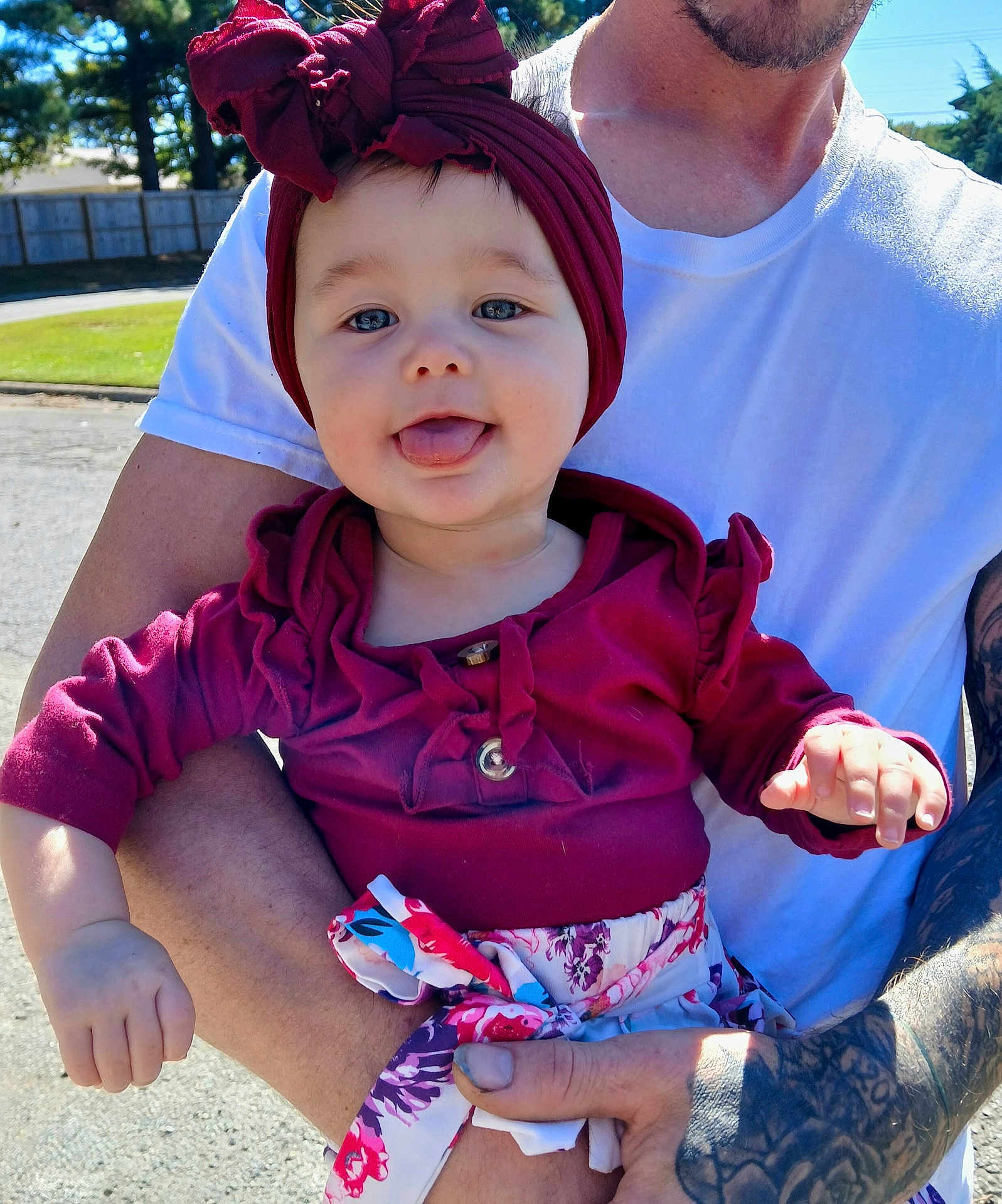 Hileigh is registered to the contest to win money with this photo: baby, child, person, adult, tattoo, outdoor, sunlight, clothing, headwrap, smiling, tongue_out, holding, arm, white_tshirt, fence, trees, nature, happy, casual, portrait