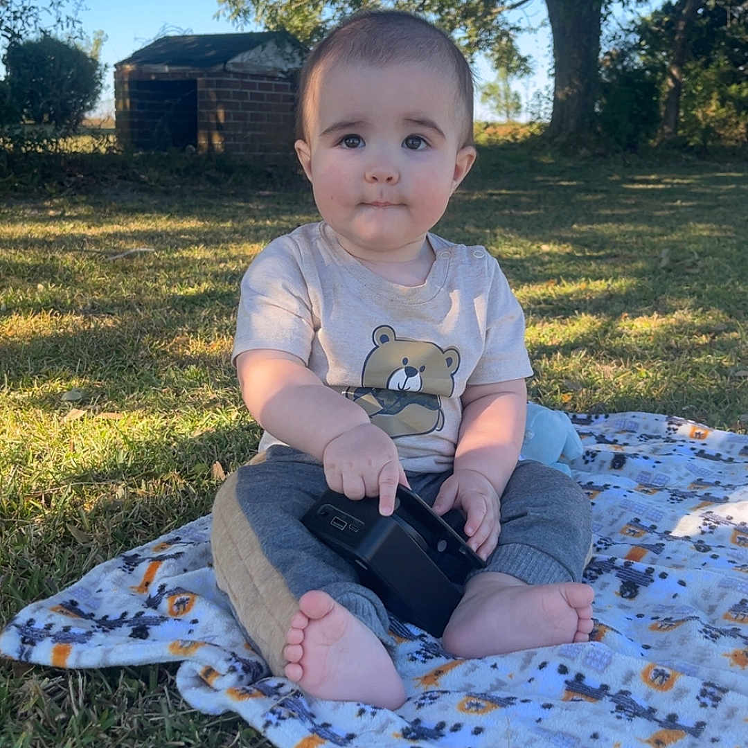 Cy joined the competition — help win amazing prizes! baby, barefoot, blanket, candid, child, clothing, curious, daylight, device, expression, face, grass, nature, outdoor, portrait, sitting, sunlight, toddler, toy, tree