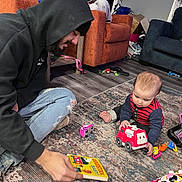 Cy is registered to the contest to win money with this photo: baby, man, adult, indoor, rug, toy, fire_truck, book, hoodie, jeans, floor, carpet, playing, child, beard, sitting, casual_clothing, home, interaction, learning