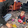 baby, man, adult, indoor, rug, toy, fire_truck, book, hoodie, jeans, floor, carpet, playing, child, beard, sitting, casual_clothing, home, interaction, learning